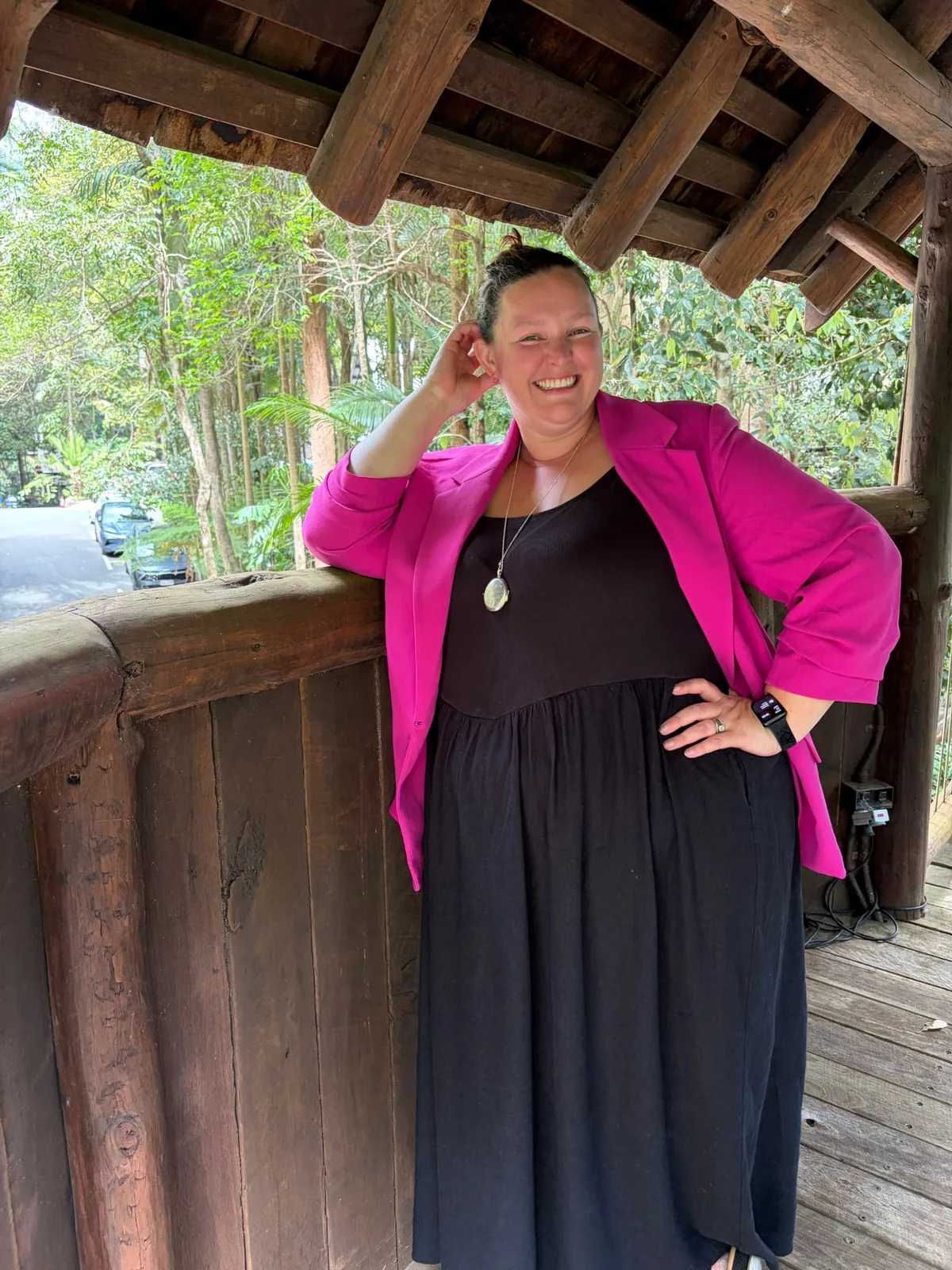 Rachel Smith laughing in a magenta blazer and black dress, leaning on a timber railing among rainforest trees