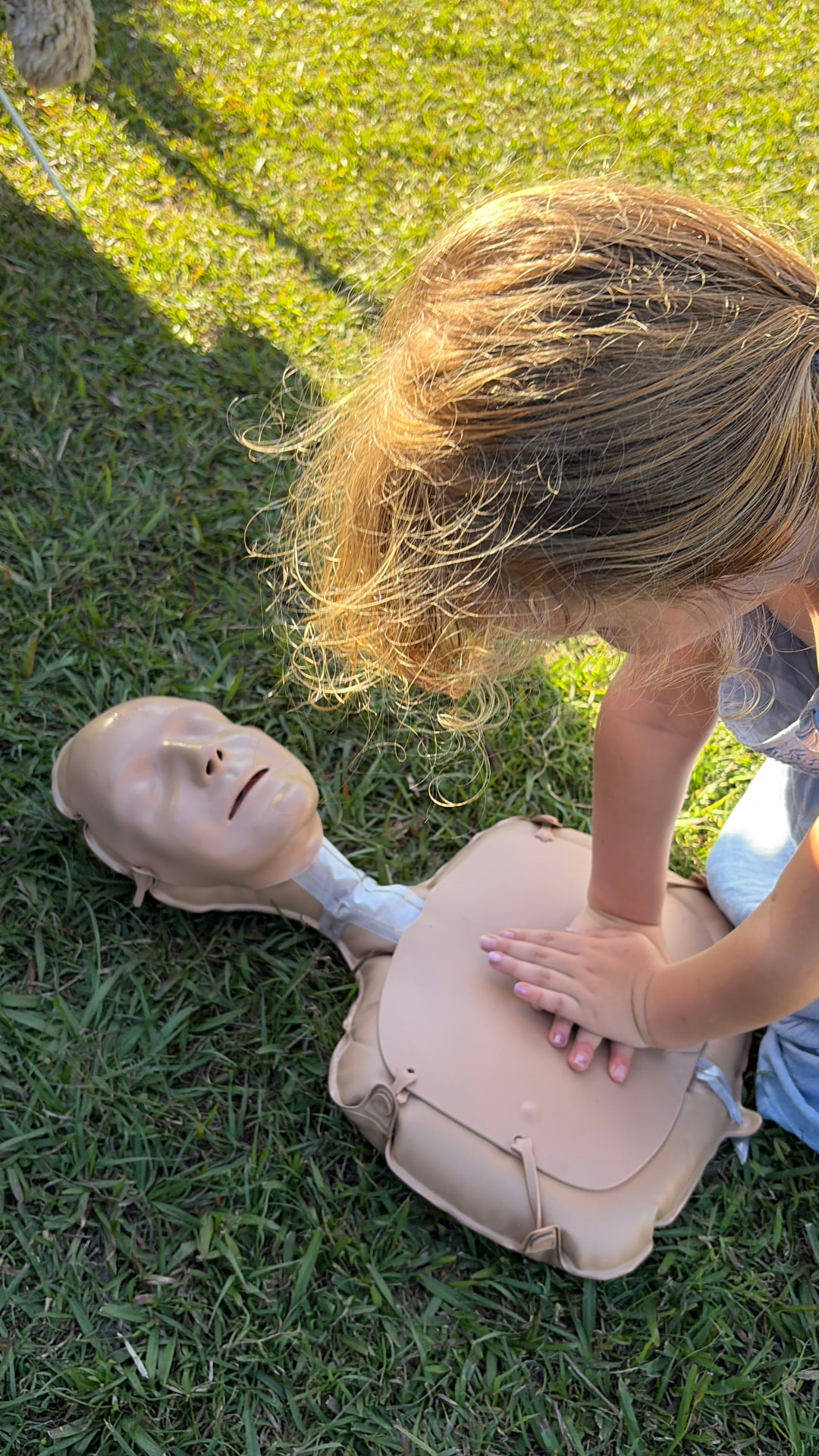 Addy kneeling on the grass with both hands placed on the chest of a CPR training manikin, practising compressions