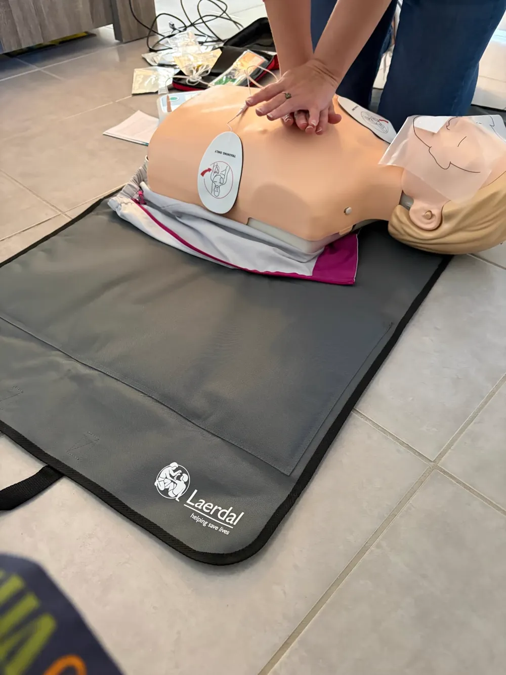 A First Aid 2 U learner giving chest compressions to an adult CPR manikin with a defibrillator training pad in place on a Laerdal training mat.