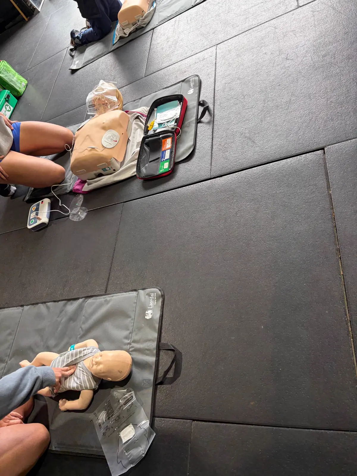First Aid 2 U training setup with an adult CPR manikin connected to a training defibrillator, an open first aid kit, and an infant manikin on a mat nearby.