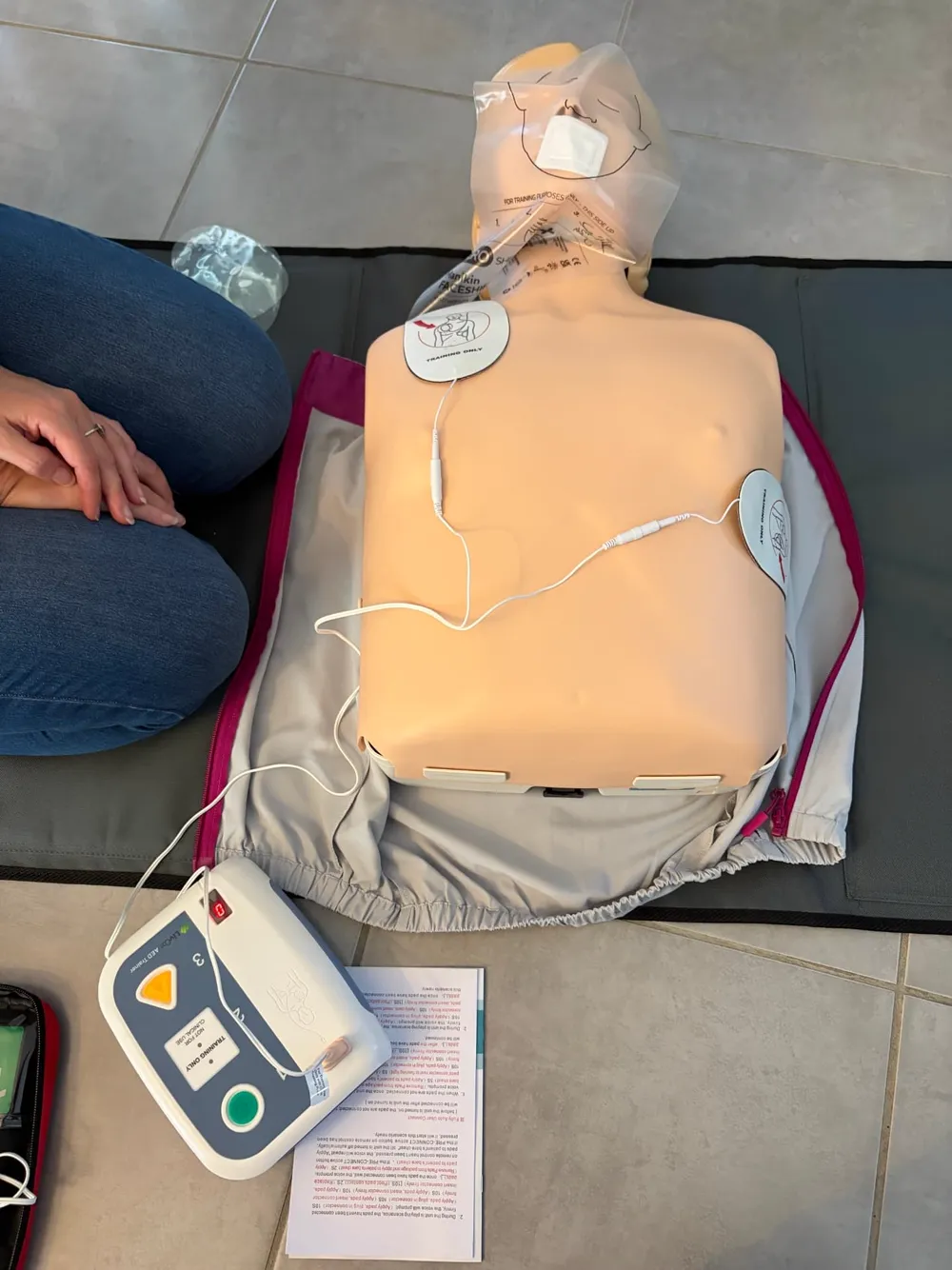 An AED trainer with its pads attached to an adult CPR manikin on a training mat, with a learner kneeling alongside during a First Aid 2 U session.