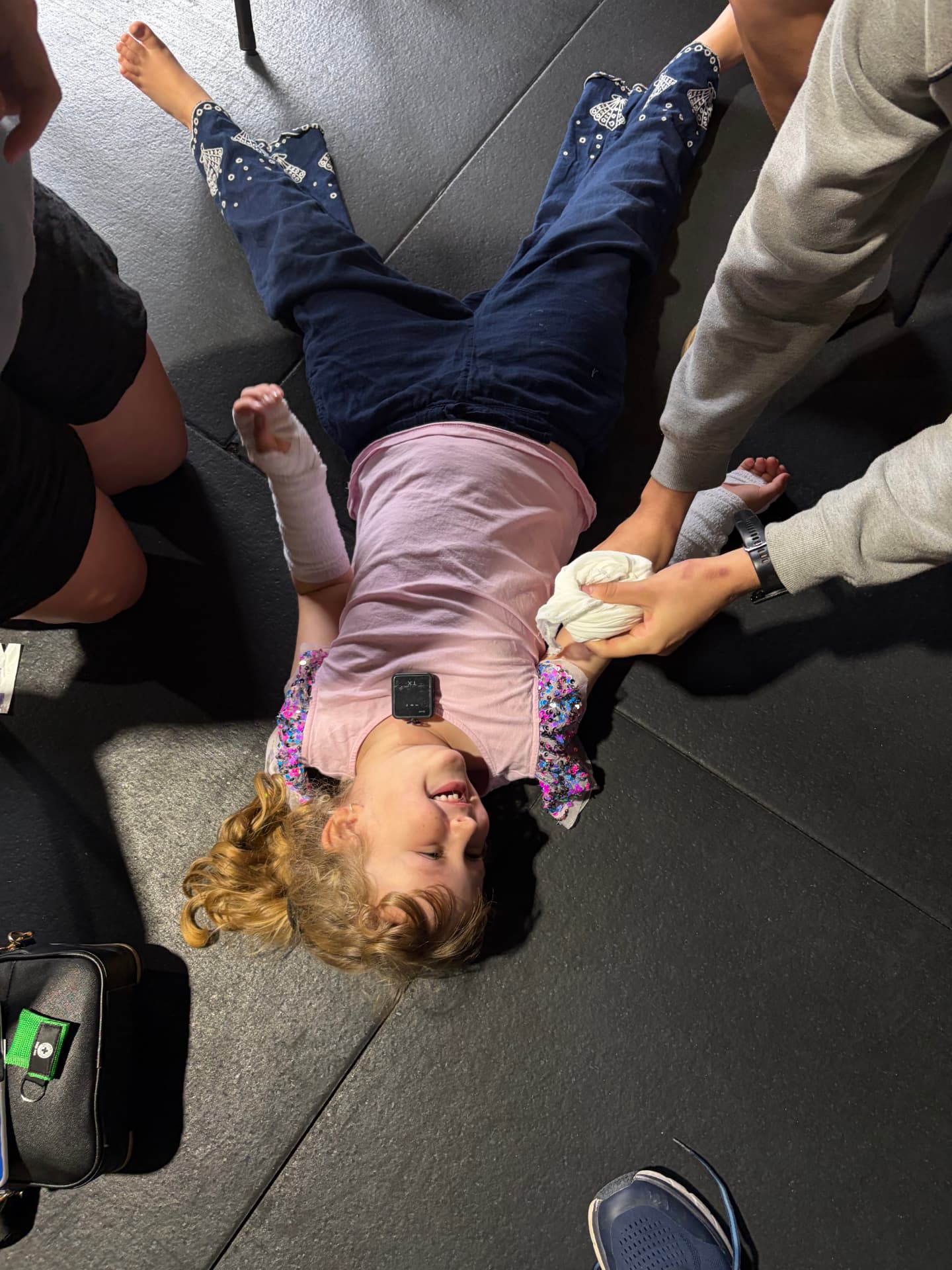 Addy lying on a mat, smiling, with bandages on her arm and foot while an adult practises bandaging her hand