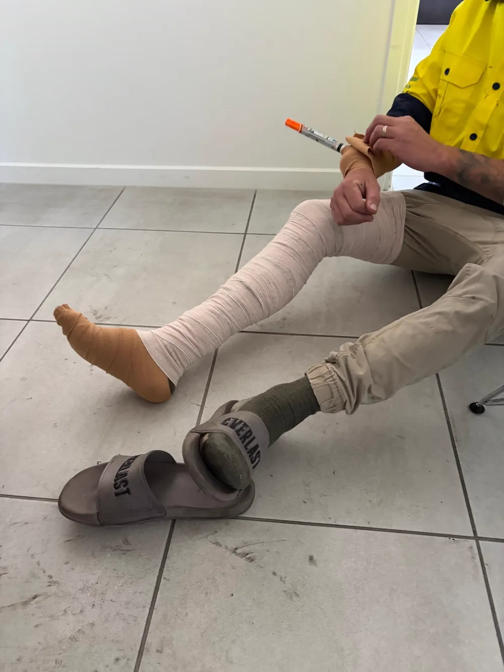 A First Aid 2 U practice scenario with a learner's leg, foot and arm wrapped in bandages while they hold an adrenaline auto-injector trainer.