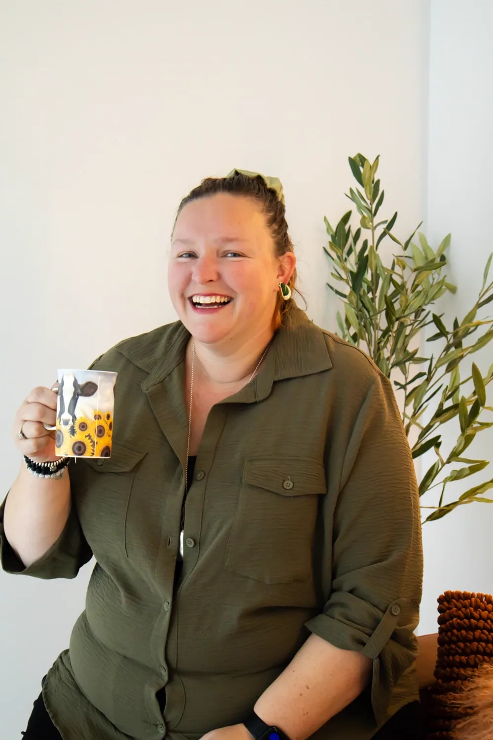 Rachel Smith laughing while holding a mug of coffee, wearing an olive shirt beside an olive plant