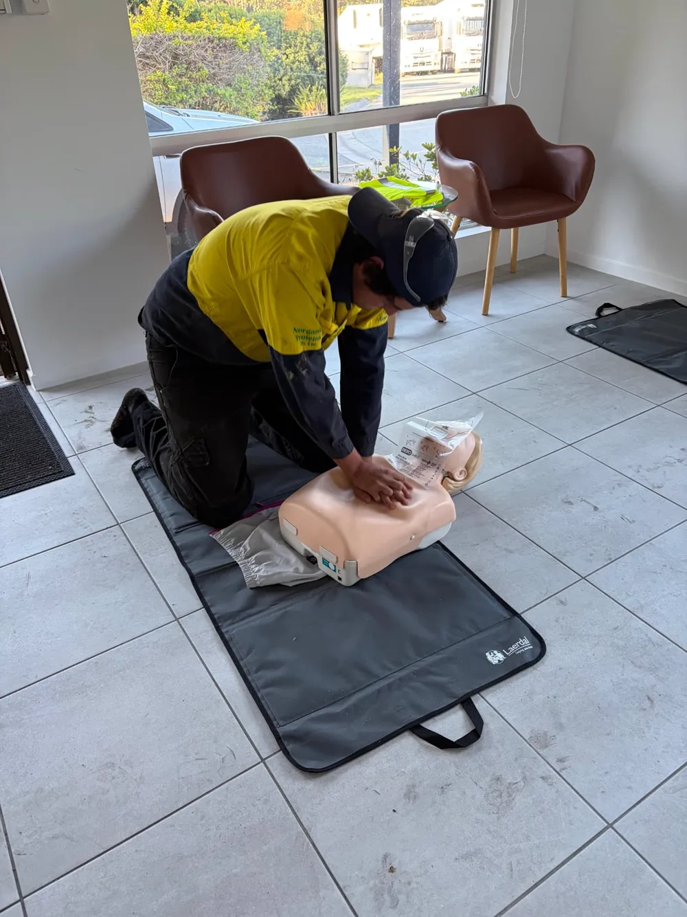 A learner in a high-visibility work shirt kneeling on a training mat and performing chest compressions on an adult CPR manikin.