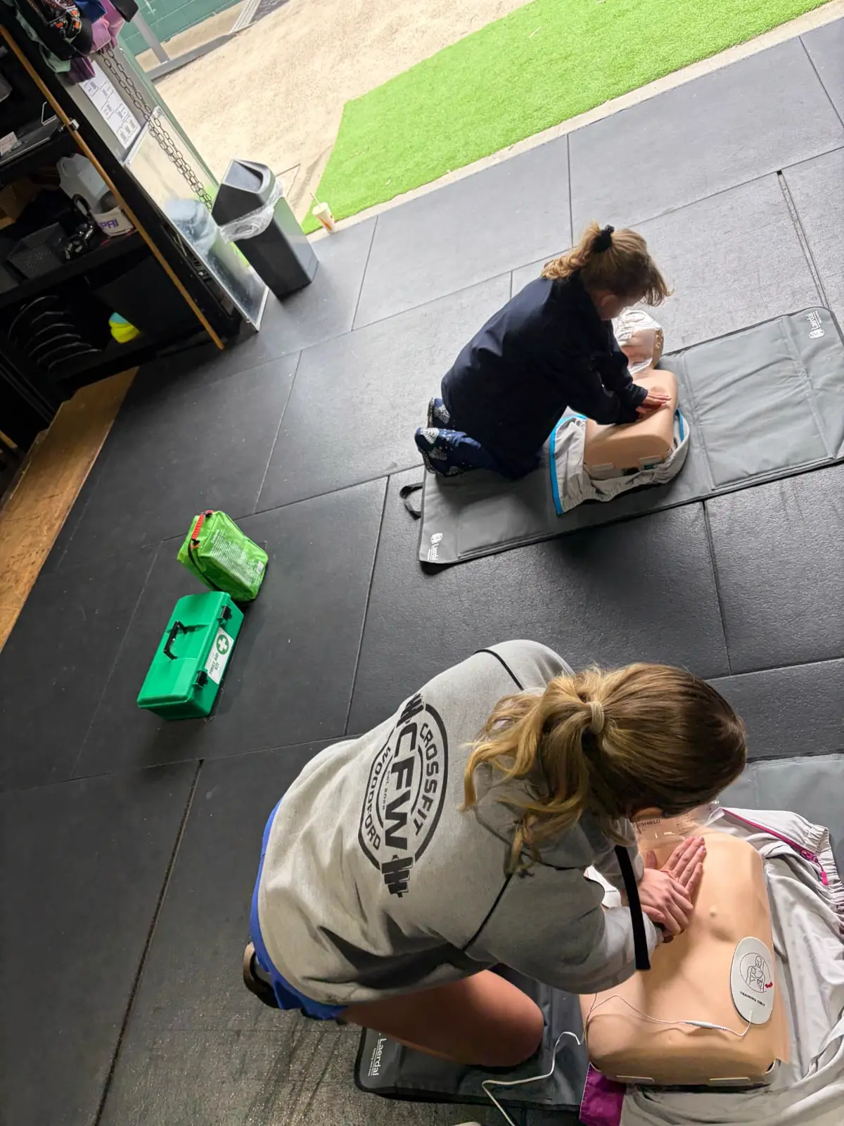 Two First Aid 2 U learners kneeling on training mats practising chest compressions on adult CPR manikins, with first aid kits beside them.