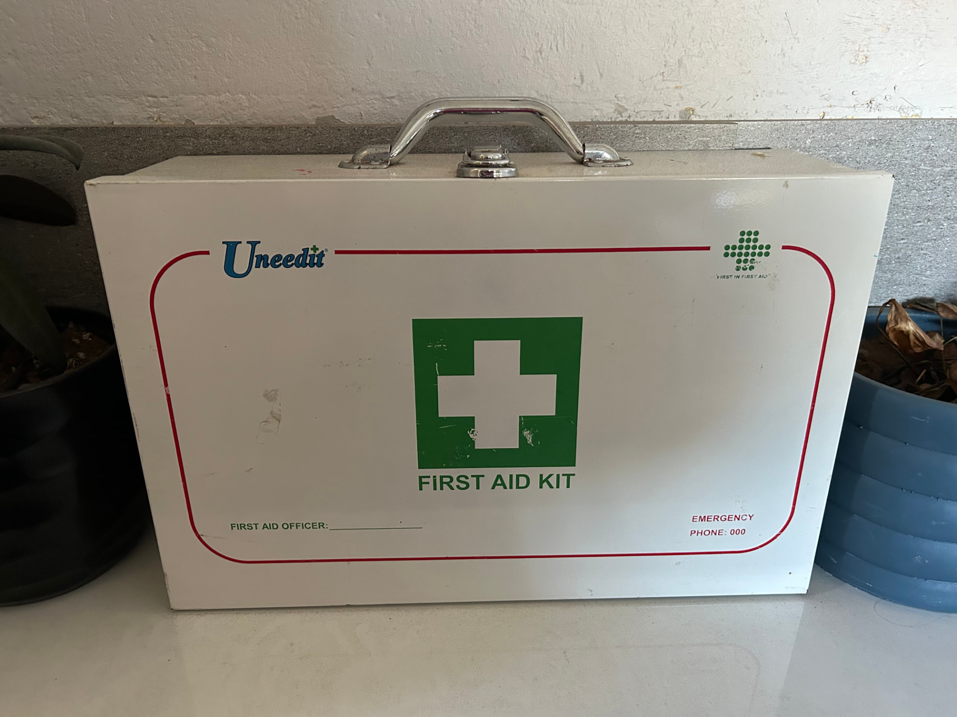 A white metal first aid kit with a green cross and Emergency phone 000 printed on the front